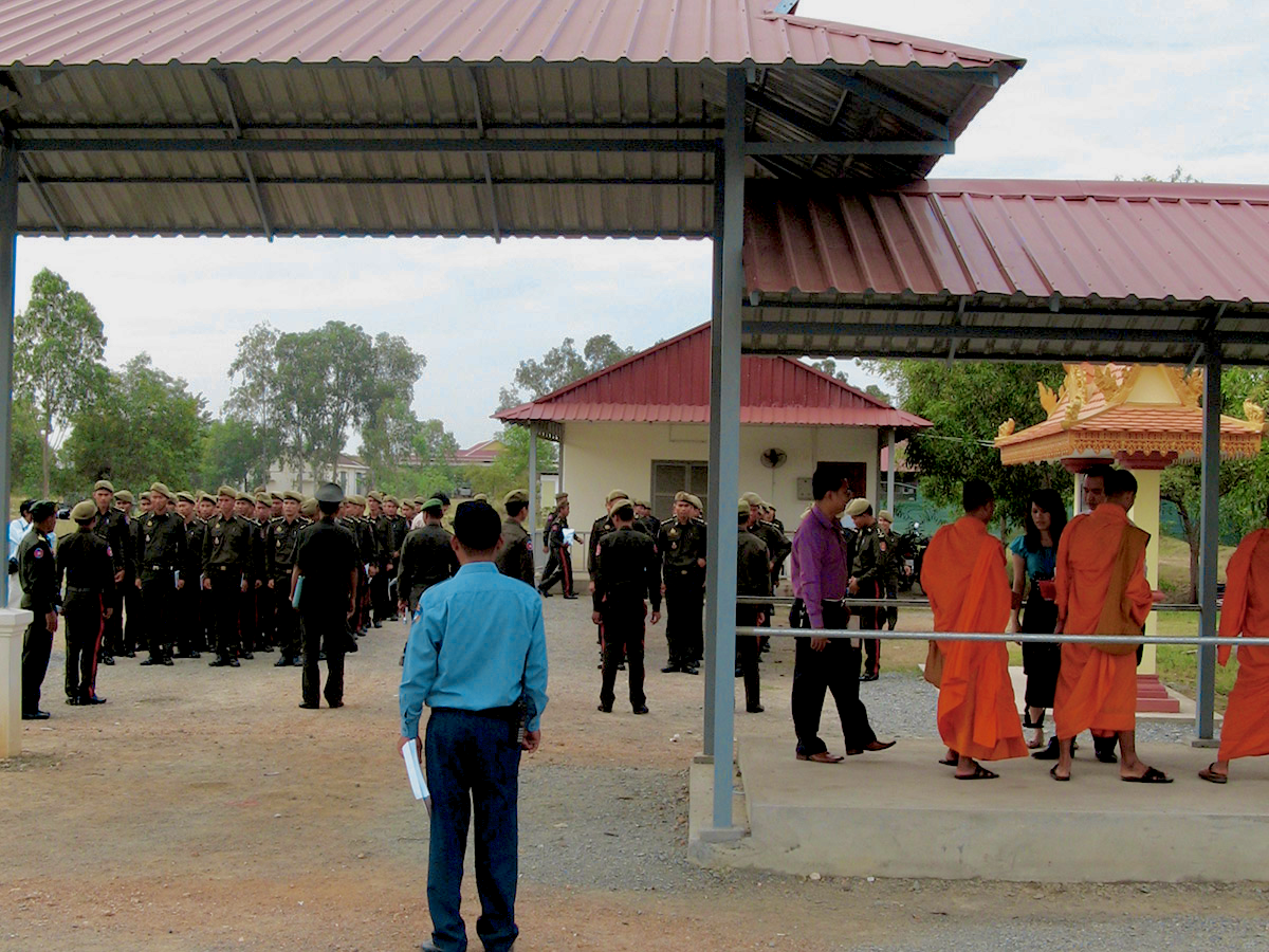 The image shows a group of men standing in front of a building, under a covered walkway. The men are dressed in military uniforms and civilian clothes. There are also a Buddhist monks, standing in the foreground under the cover.
