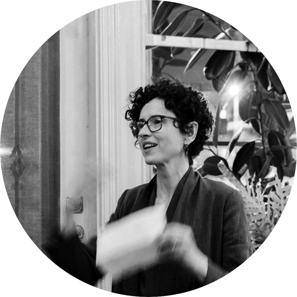 A black and white image of a woman talking. The woman has short, dark, curly hair and is wearing glasses and a suit jacket. 