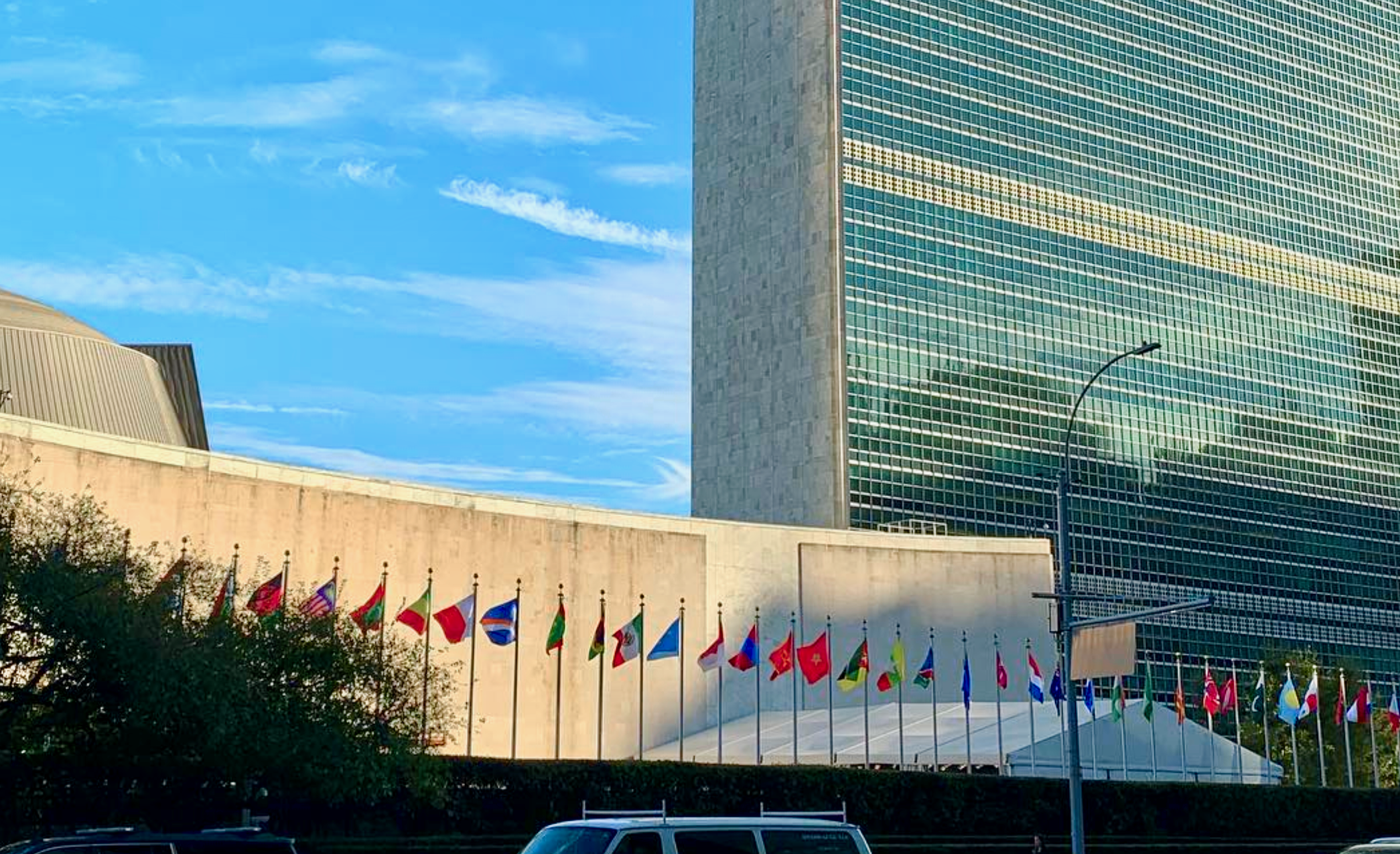 An image showing over 30 flag poles hosting flags of global countries spanning the width of the image. In the foreground are parked cars, and in the background is a glass clad skyscraper.